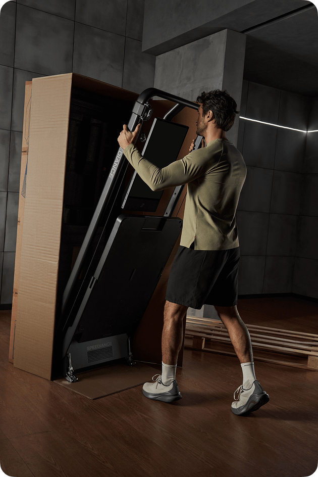 Person exercising on a fitness machine with another person assisting them, in a home setting with a view of trees.