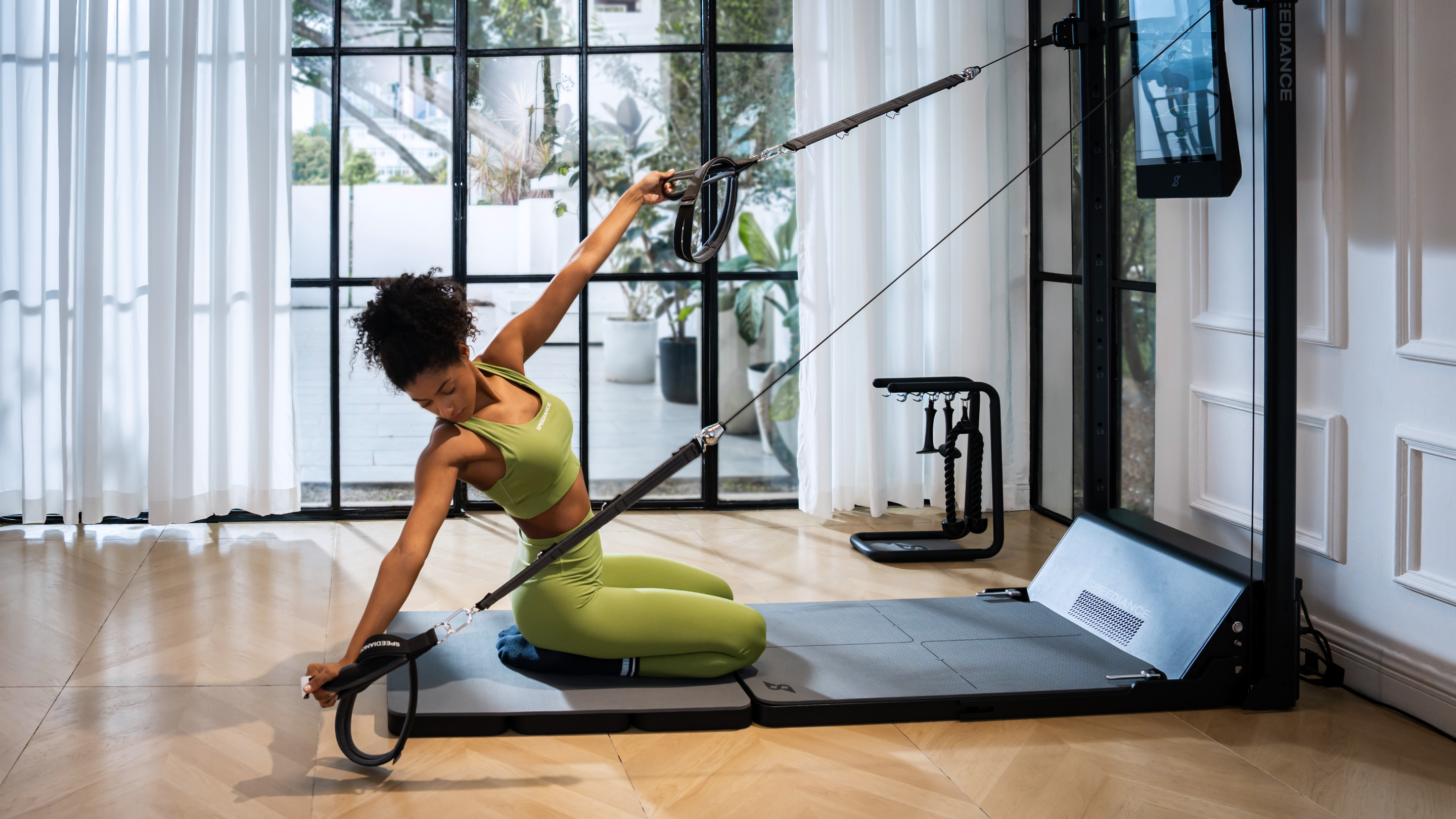A woman in a green sports bra and leggings performs a Pilates exercise on a reformer machine in a bright room with large windows and white curtains.