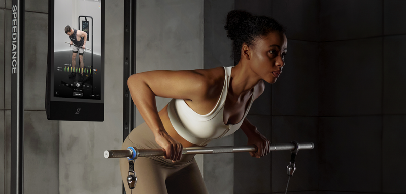 Woman exercising with a barbell in a gym setting, using a Speeddance machine.