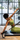 A woman in a lime green sports bra and matching leggings exercises on a reformer machine in a bright gym with large windows.