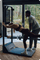 Person exercising on a fitness machine with another person assisting them, in a home setting with a view of trees.