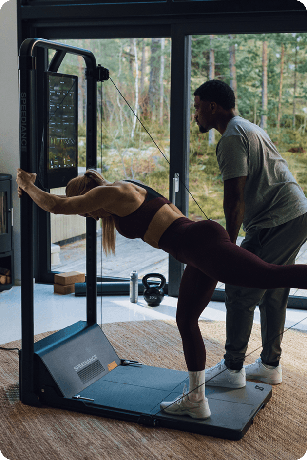 Person exercising on a fitness machine with another person assisting them, in a home setting with a view of trees.