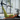 A woman in a green sports bra and leggings lies on a Pilates reformer machine, extending one leg upward into the attachment. The setting is a bright, modern fitness studio with large windows and white curtains.