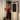 A woman stands using a black home exercise machine with a touch screen display, holding resistance bands. She is wearing a rust-red tank top and black shorts in a bright indoor room with white curtains and paneled walls.